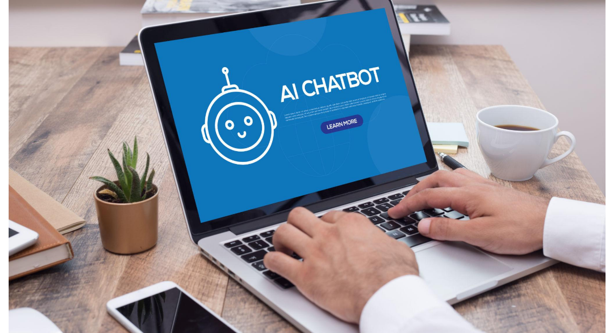 ノートパソコンの画面に『AI CHATBOT』と表示されたインターフェースを開き、机の上でキーボードを操作する人物の手元。横にはスマートフォンやコーヒーカップが置かれている。