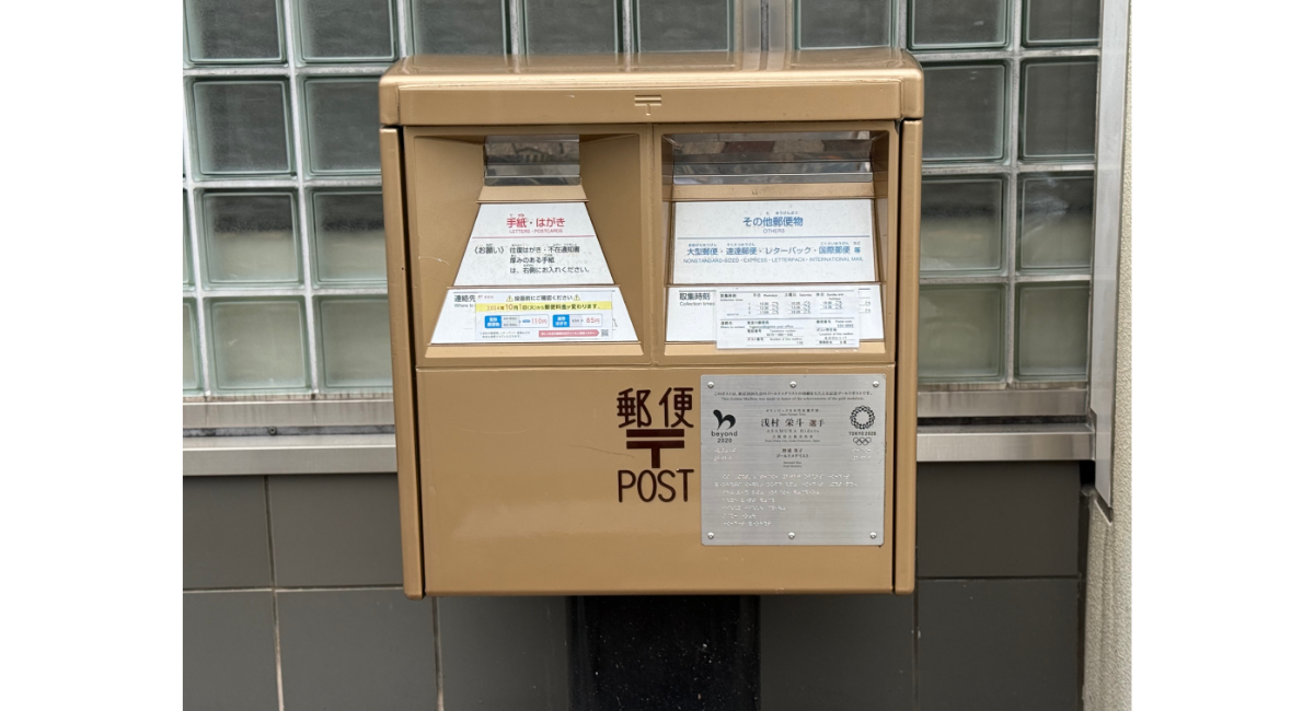 金色に塗装された日本の郵便ポストのクローズアップ。前面に『郵便 POST』の表記と東京オリンピック関連の記念プレートが取り付けられている様子。