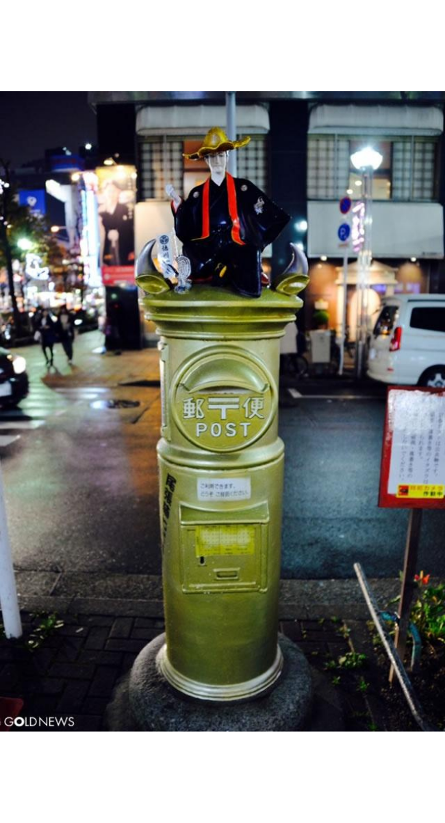 夜の街角に設置された金色の丸型郵便ポストの上に、和装で金色の帽子をかぶった人形が飾られている様子。周囲にはネオンや車が見える都市風景。