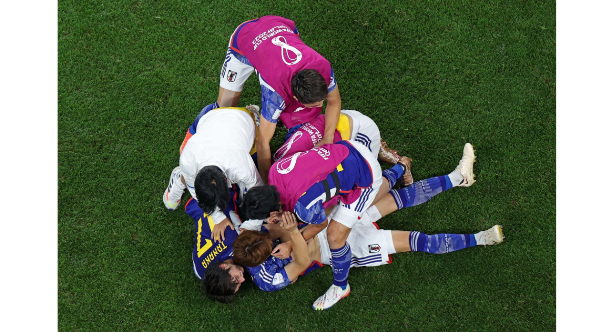 サッカー日本代表の選手たちがピッチ上で重なり合い、得点後の喜びを分かち合っているシーン。芝生の上で複数の選手が集まり、チームの一体感と熱量が伝わる瞬間を捉えた画像。