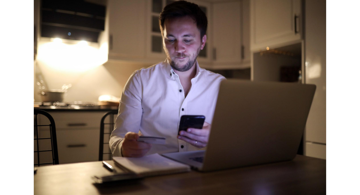 夜の室内でノートパソコンの前に座り、スマートフォンとクレジットカードを手に操作する男性の様子。オンライン取引やネット賭博、金銭管理のリスクをイメージさせる場面。
