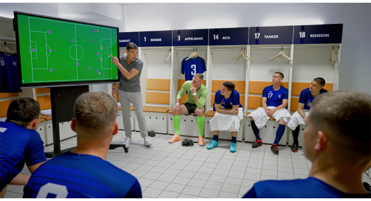 ロッカールームでコーチが戦術ボードを使って試合の戦略を説明し、選手たちが座って聞いているサッカーチームのミーティングの様子。