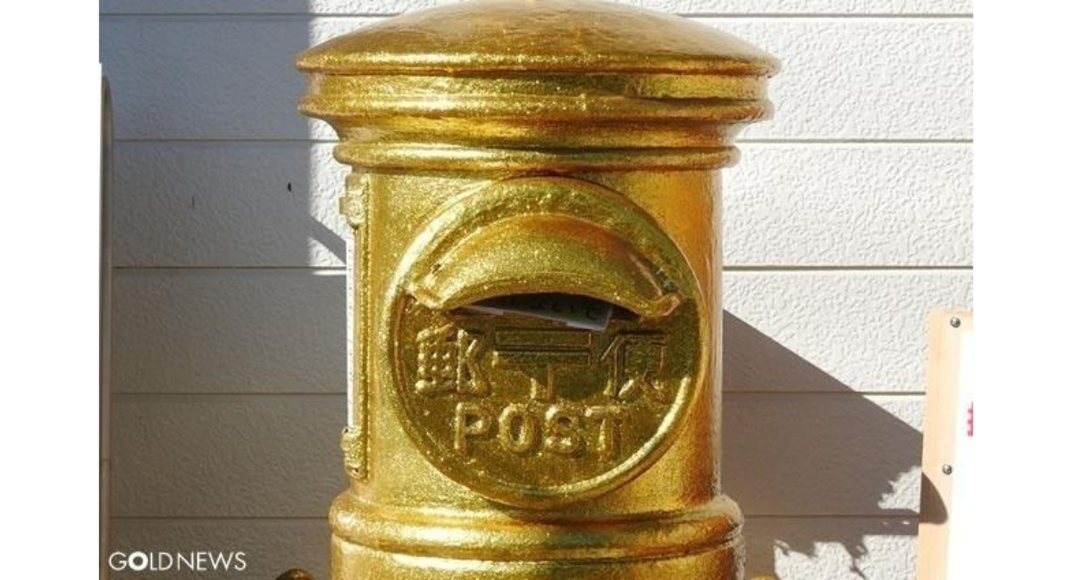 金色に塗られたレトロな丸型郵便ポストの正面。投函口の下に『郵便 POST』の浮き彫り文字があり、壁際に設置された様子。