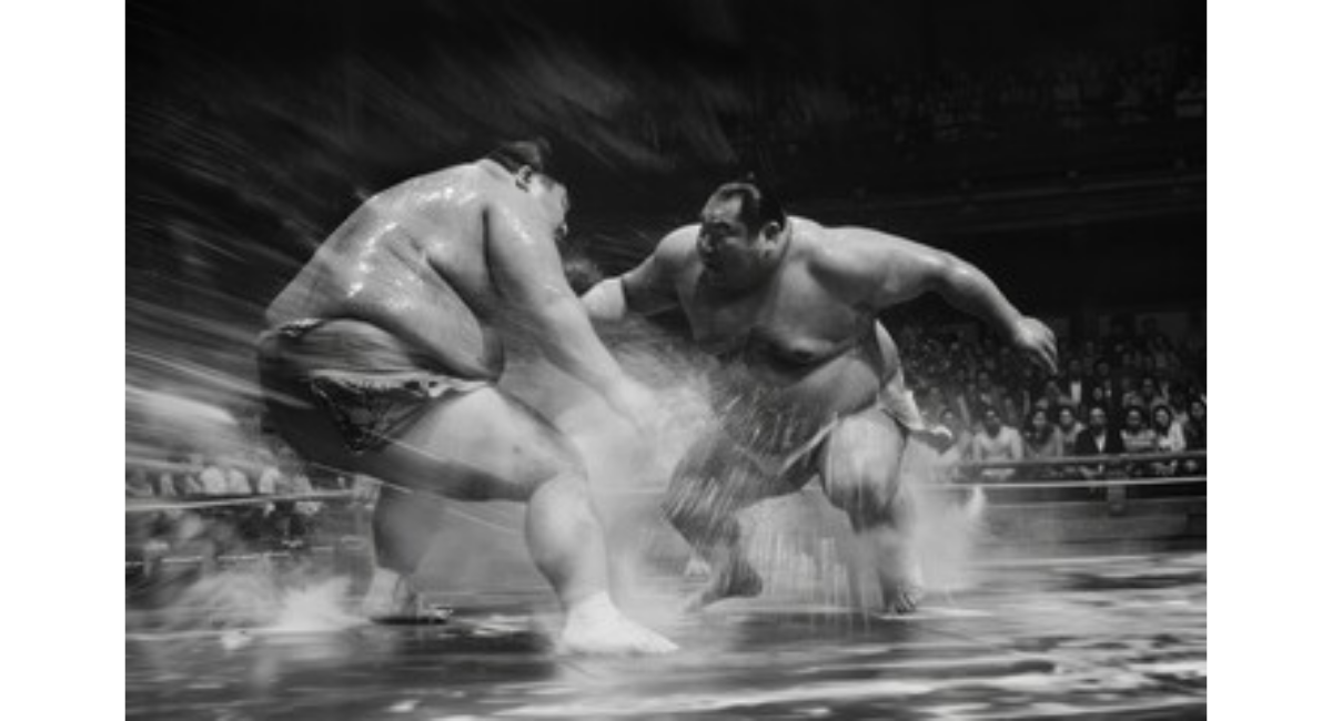 力士同士が立ち合いで激しくぶつかる瞬間を捉えたモノクロ画像。土俵上に砂煙が舞い、迫力ある相撲の攻防を表現したシーン。