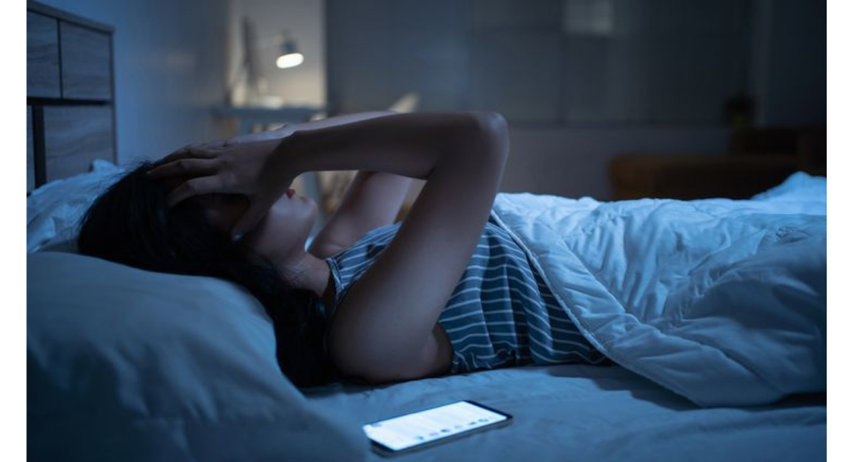 夜のベッドでスマートフォンの光を浴びながら眠れずに悩む女性の様子を表した、SNS利用による睡眠不足のイメージ画像